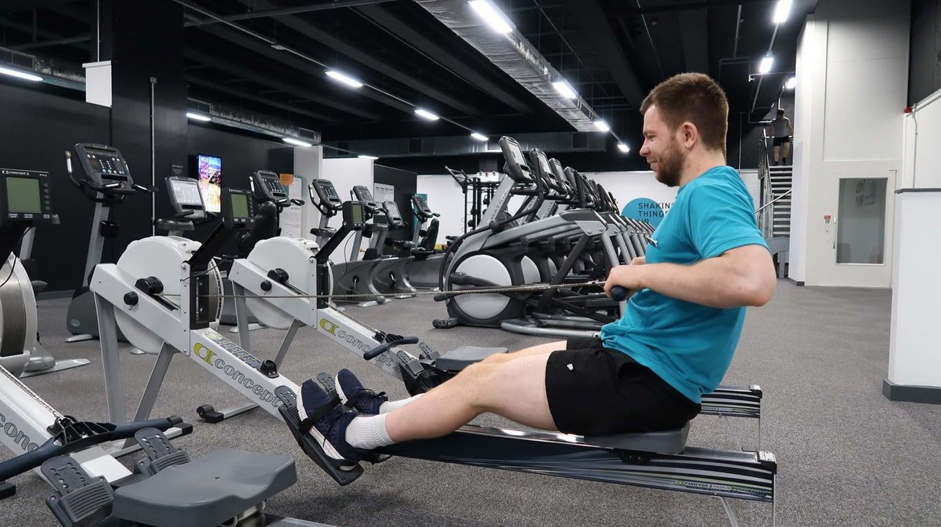 Man in blue shirt using rowing machine in gym with exercise bikes and treadmills nearby