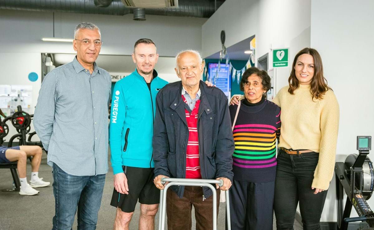 Five people standing together in a gym, one elderly man using a walker.