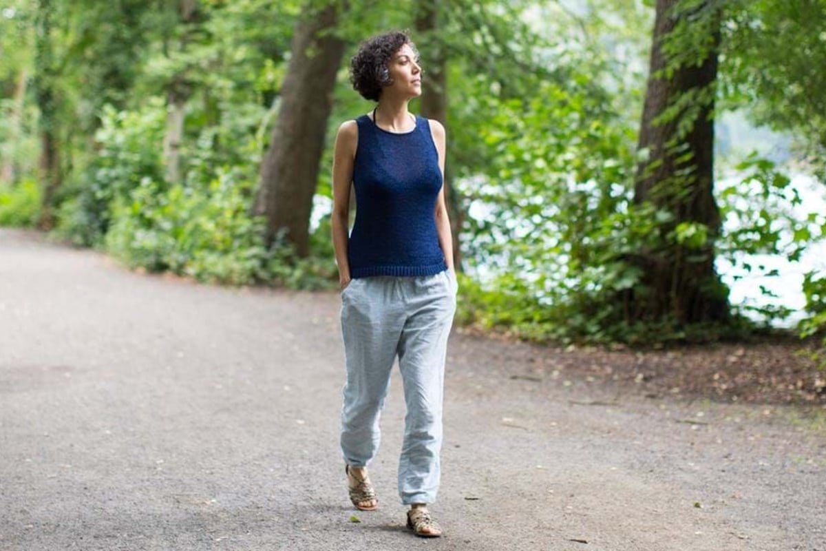 Woman in blue sleeveless top and light trousers walking on a forest path with hands in poc