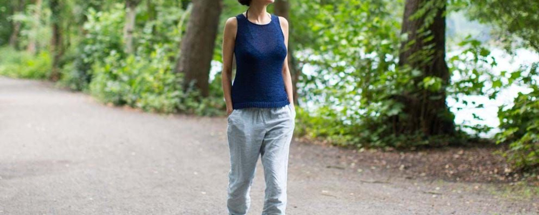 Woman in blue sleeveless top and light trousers walking on a forest path with hands in poc