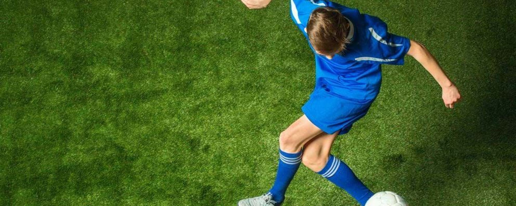 Boy in blue kit kicking a white football on green grass pitch from above