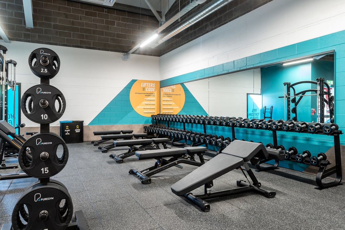 PureGym Aberdare: Weightlifting area with benches, barbell racks, weight plates, and motivational graphics on walls.