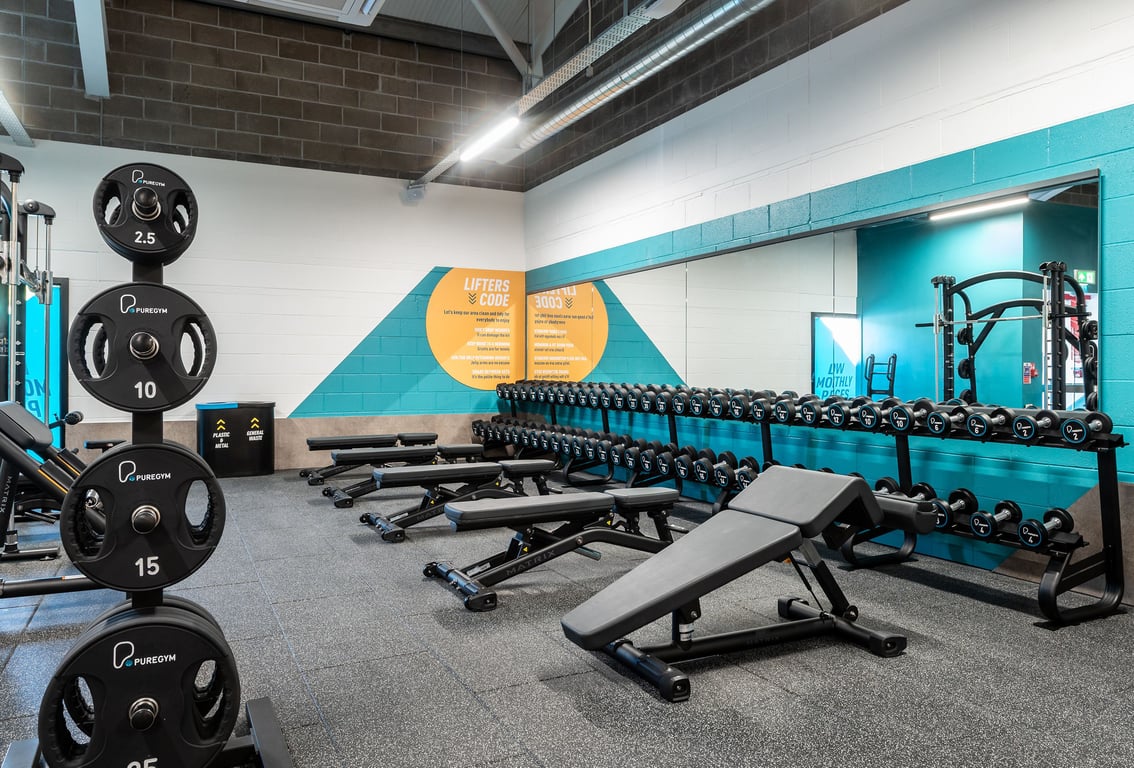 PureGym Aberdare: Weightlifting area with benches, barbell racks, weight plates, and motivational graphics on walls.
