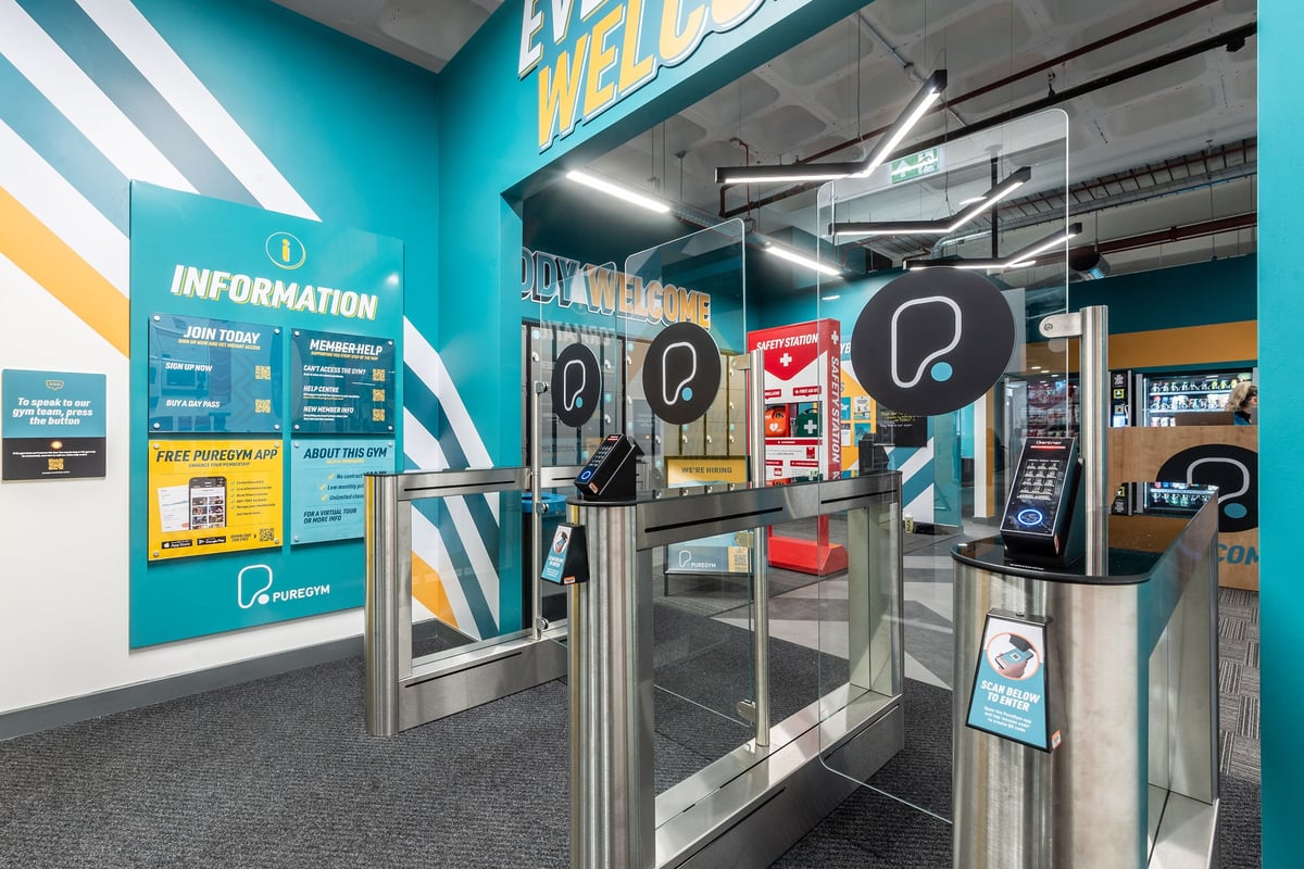 PureGym London Wood Green: Check-in area with informational displays, self-service kiosks, and security gates.