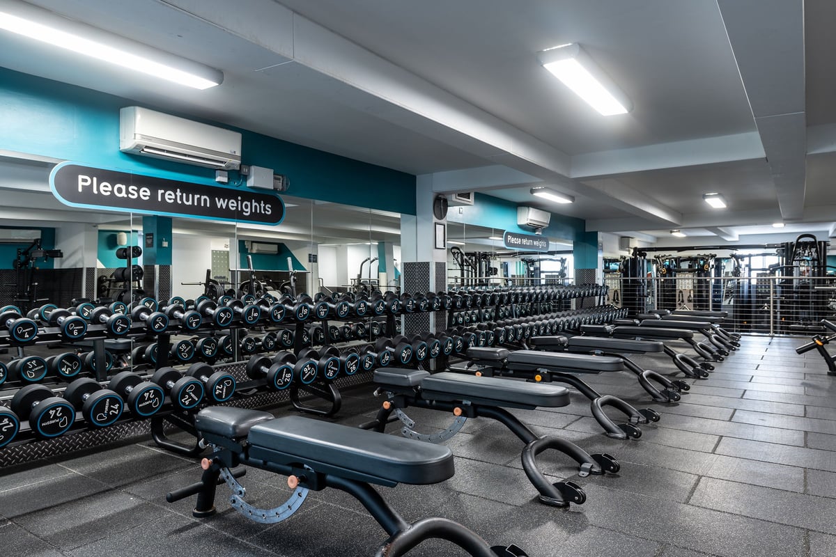 PureGym Sutton Coldfield: Rows of dumbbells, weighted bars, benches, and rubberized flooring in a teal-tinted gym interior.