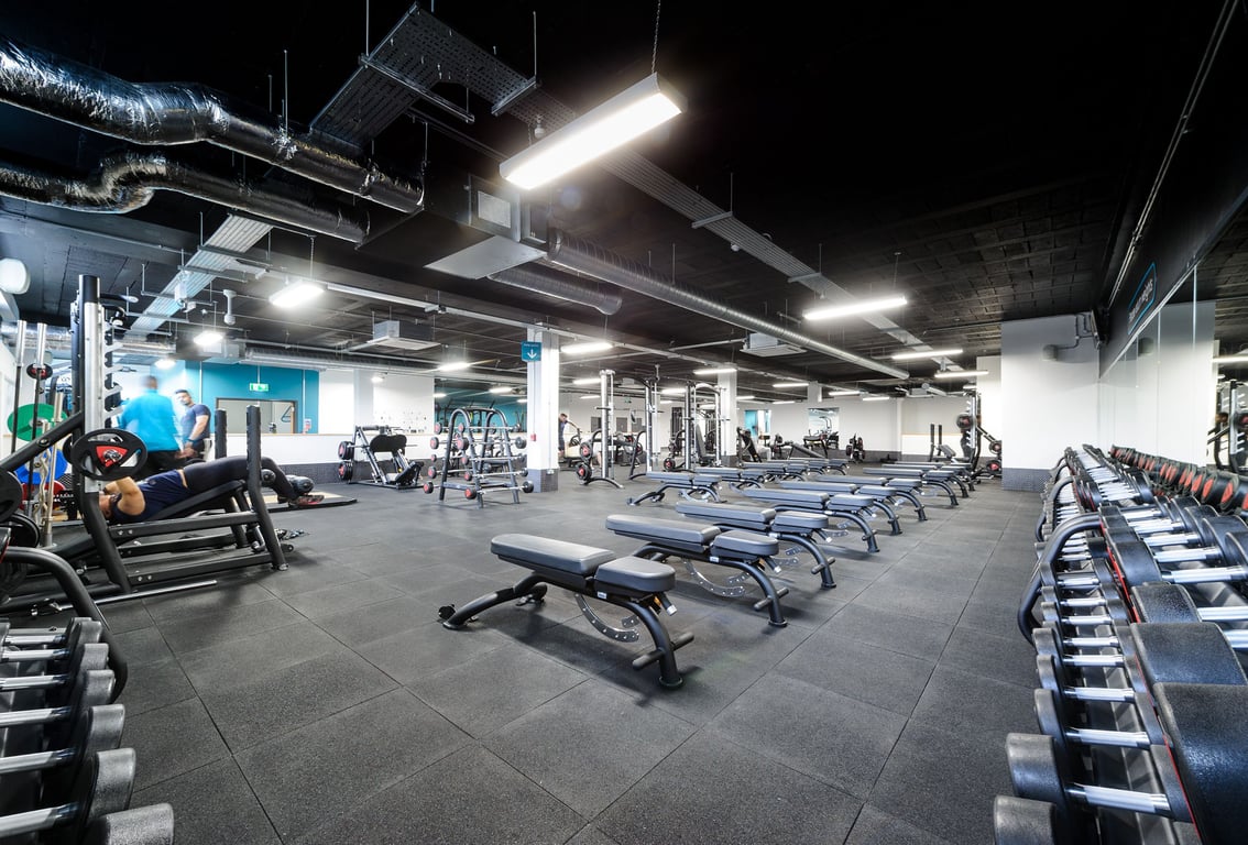 PureGym London Bromley: Weight benches, free weights, squat racks, mirrored walls, and exposed ducts in dimly-lit gym.