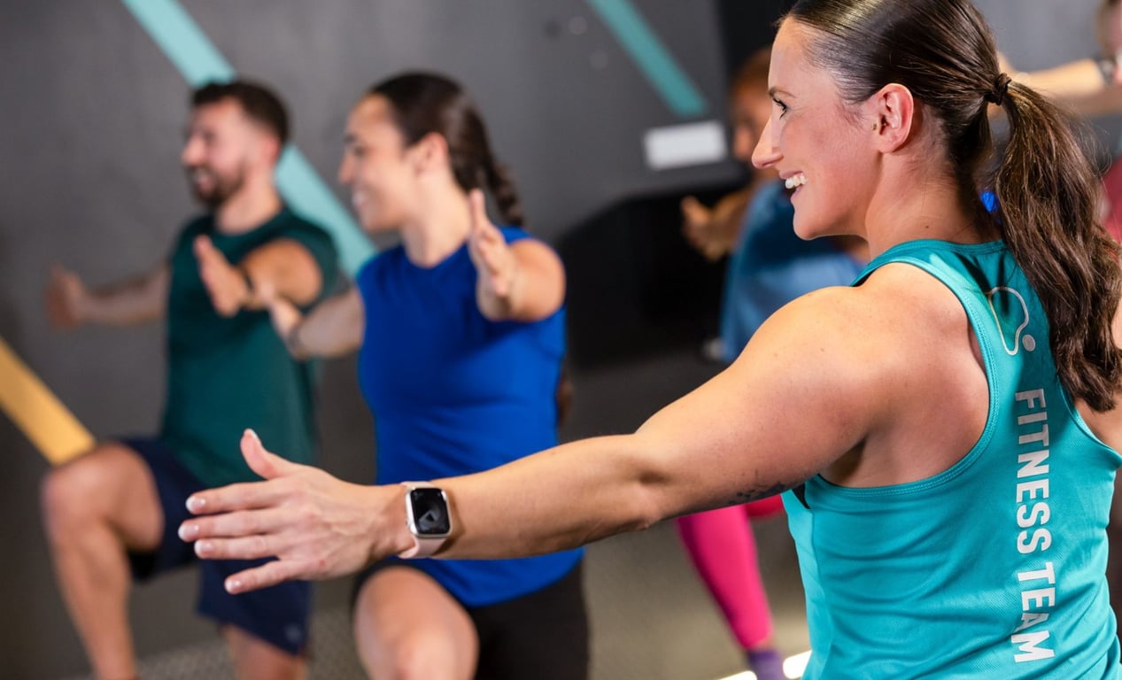 PureGym: a HIIT class lead by a female instructor stretching out her arm, with a female and a male member raising their knees in the background
