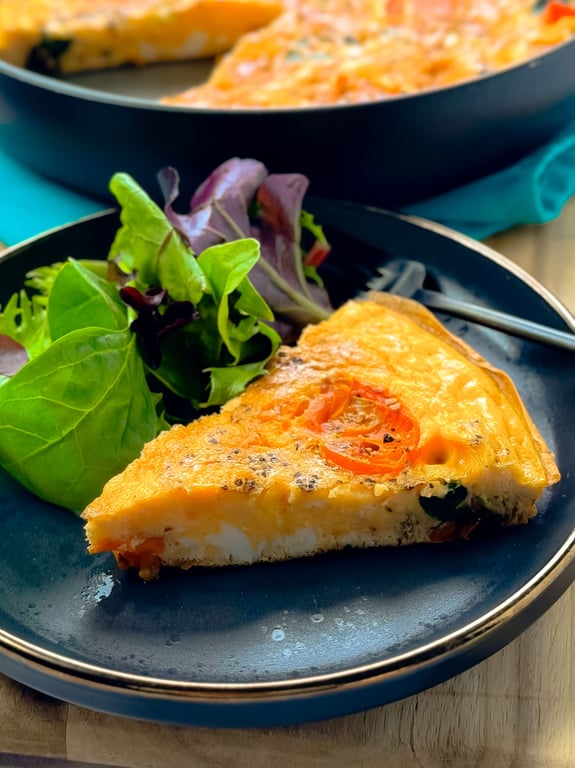Slice of vegetable frittata with salad greens on a dark plate with a fork