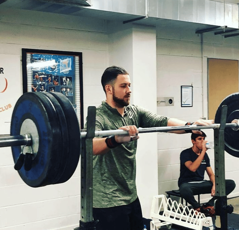 Man adjusting barbell loaded with weights in gym while another drinks water sitting down