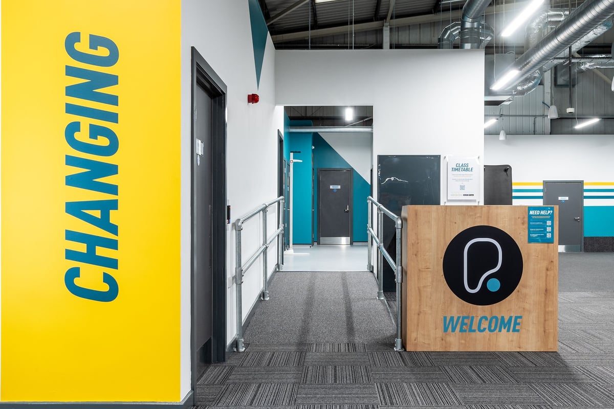 PureGym Newhaven: Long corridor, directional signage "Changing", reception desk with "Welcome" sign.