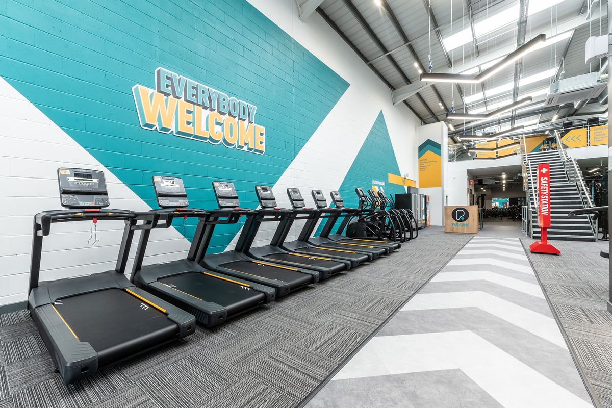 PureGym Chippenham: Rows of treadmills, turquoise accent wall with 'Everybody Welcome' signage, industrial ceiling.