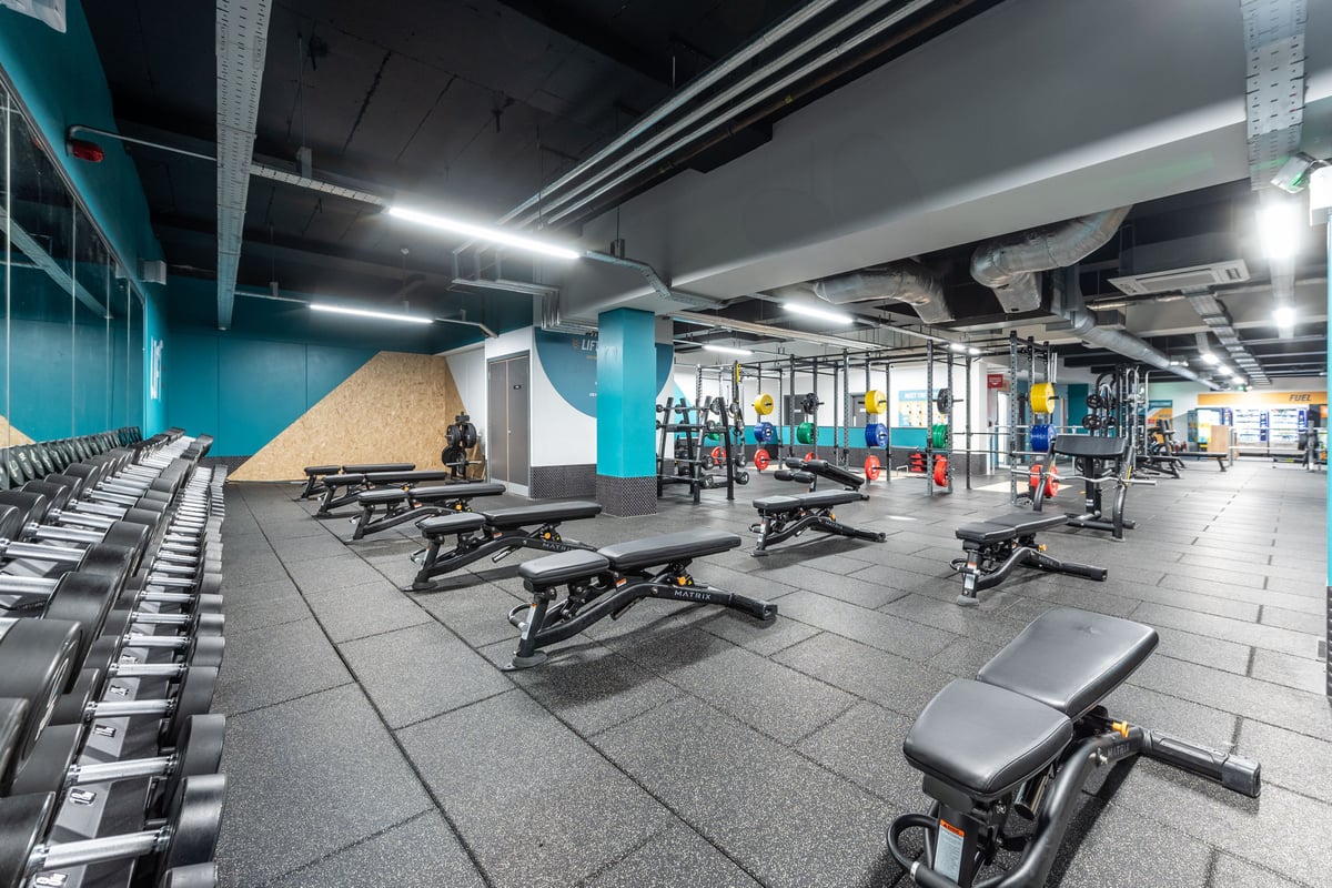 PureGym London Ilford: Weightlifting area with benches, racks, dumbbells, and vibrant teal-coloured walls.