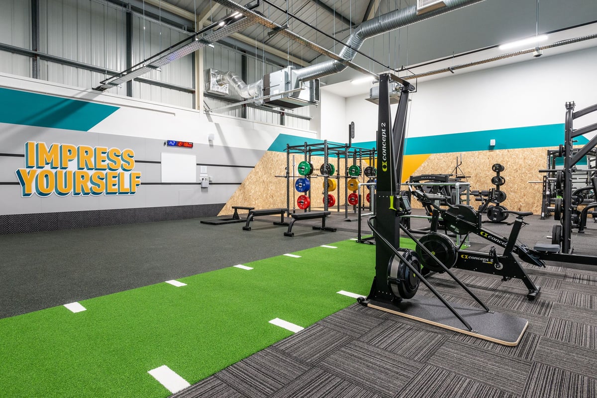 PureGym Oswestry: Squat racks, weight plates, turf area, rubberized flooring in a fitness studio with motivational branding.