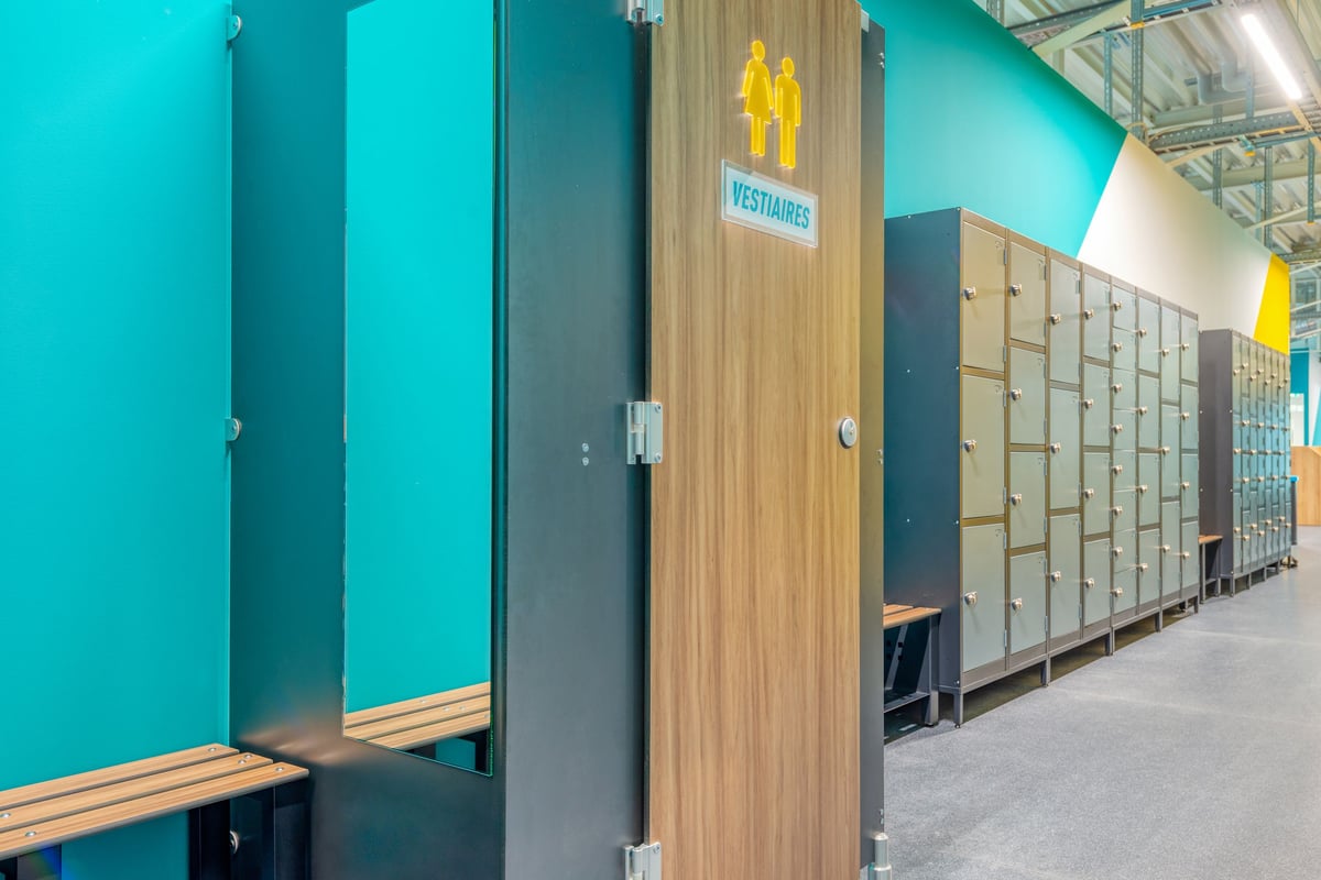 PureGym Martigny: Toilettenkabinen, Umkleideräume und Schränke säumen blaugrüne Wände mit Holzlaminattüren.