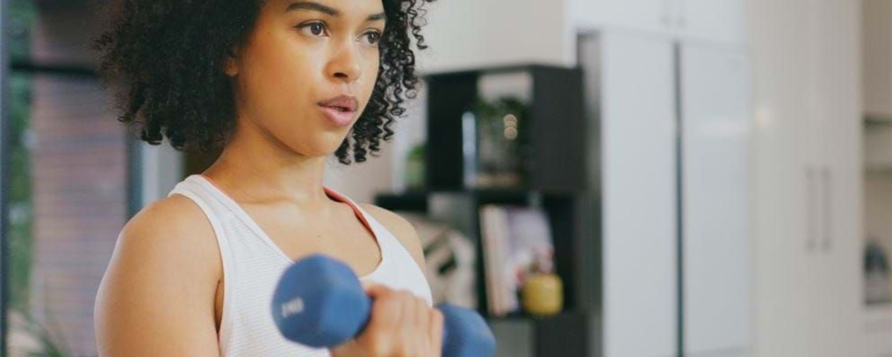 Woman lifting a blue dumbbell in a white tank top in a modern kitchen
