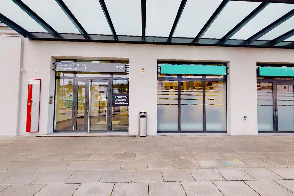 PureGym Olten: Aussenansicht eines Gebäudes mit gläsernen Eingangstüren, einem roten Stehstand und grün akzentuierten Fenstern.