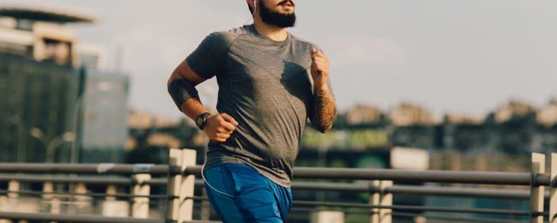 Man with beard and tattoos jogging outdoors near metal railings in cityscape background