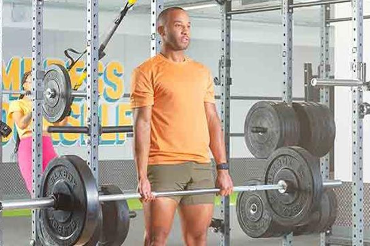 Mann im orangefarbenen Hemd hält Hantelstange in Fitnessstudio mit Gewichten und Ausrüstun
