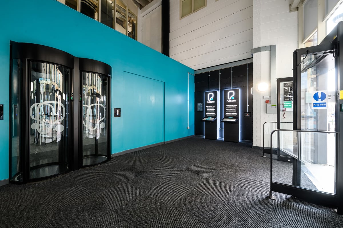 PureGym Birmingham Arcadian Centre: Revolving glass doors with graffiti-style imprints, teal walls, carpet floor.