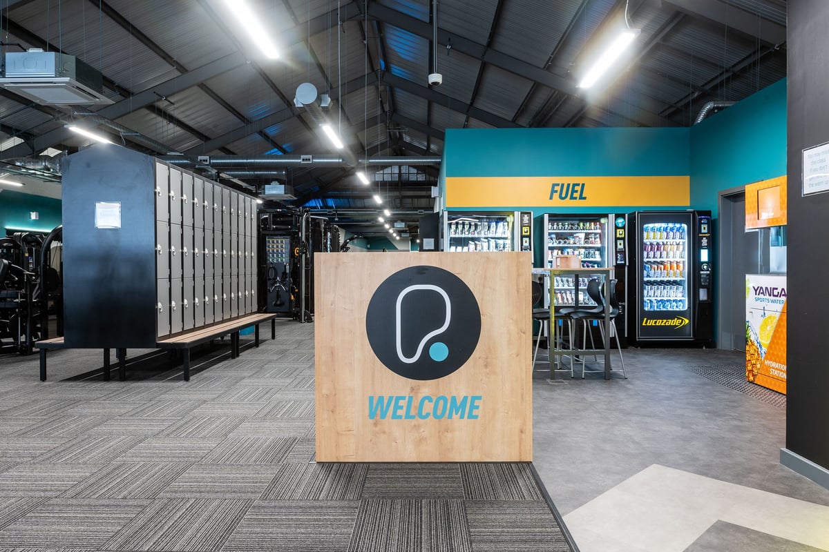 PureGym Derby Meteor Centre: Entrance area with a welcome podium, vending machines with food and beverage, and lockers.