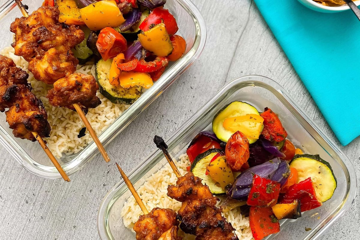 Two glass containers with grilled chicken skewers, rice, and mixed roasted vegetables