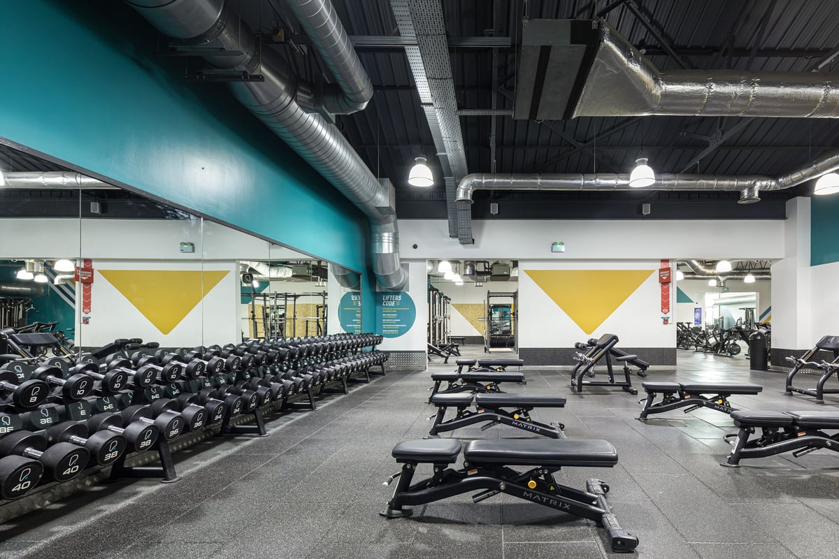 PureGym Dudley Tipton: Rows of weight benches, free weights, and mirrors line a gym's floor with teal and yellow accents.
