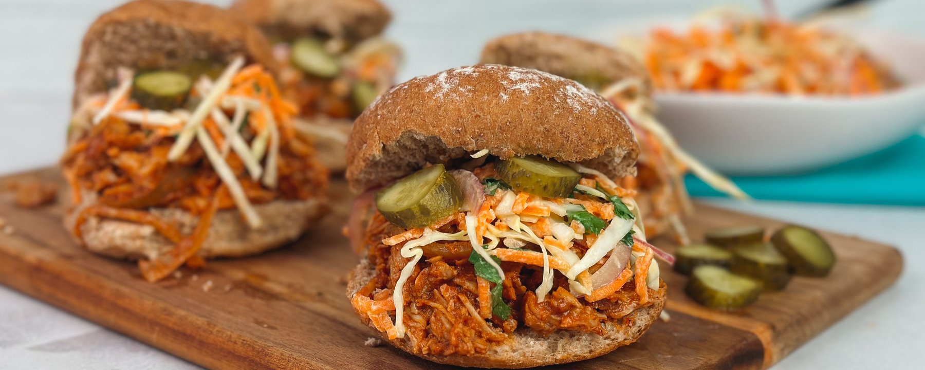 Pulled pork burgers with coleslaw and pickles on a wooden board beside a bowl of slaw