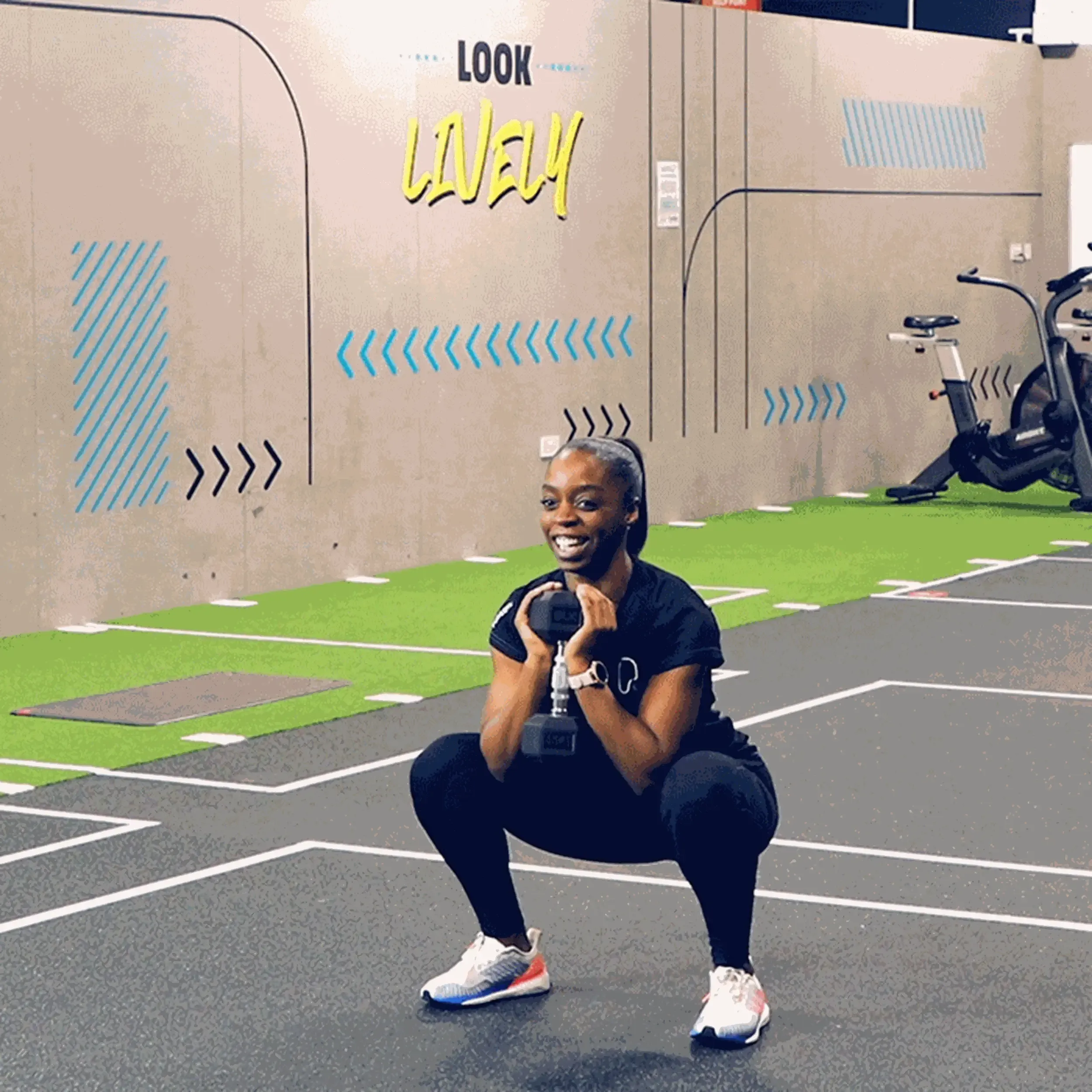 Woman squatting with a dumbbell in a gym with "Look Lively" on the wall behind her