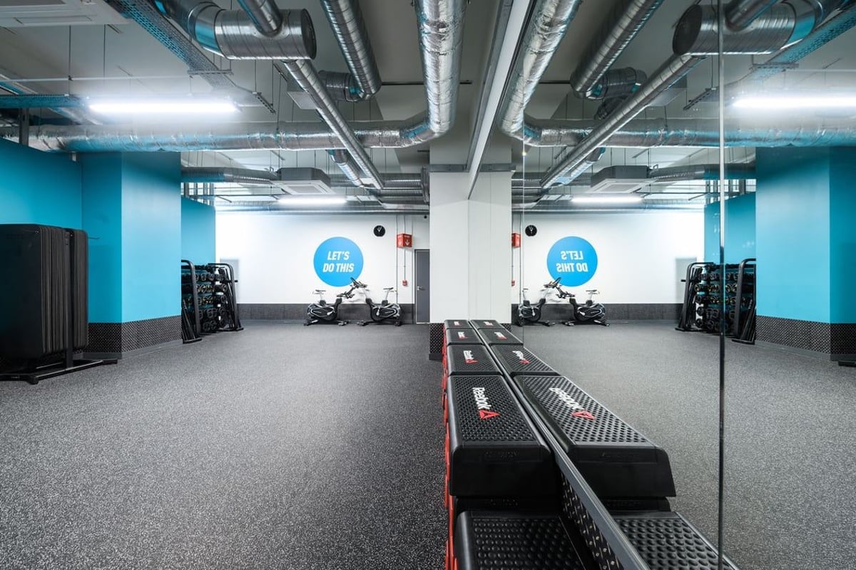 PureGym London Whitechapel: Blue room with weight racks, free weights, mirrors, and exposed overhead pipes/ducts.