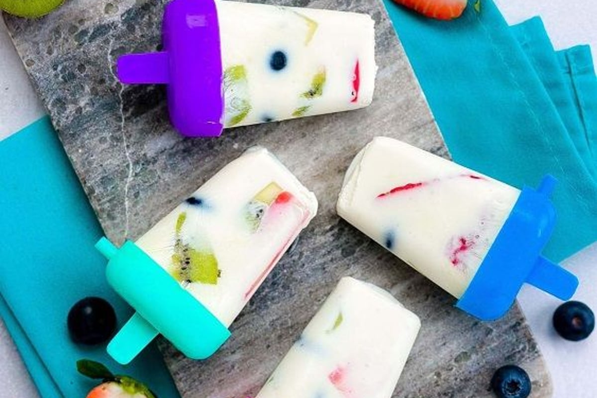 Frozen yoghurt popsicles with kiwi, strawberry, and blueberry on a marble board.