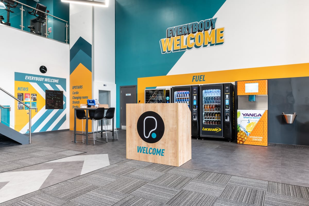 PureGym Peterborough Brotherhood Retail Park: Vending machines, welcome sign, fuel logos, teal and yellow branding on walls.