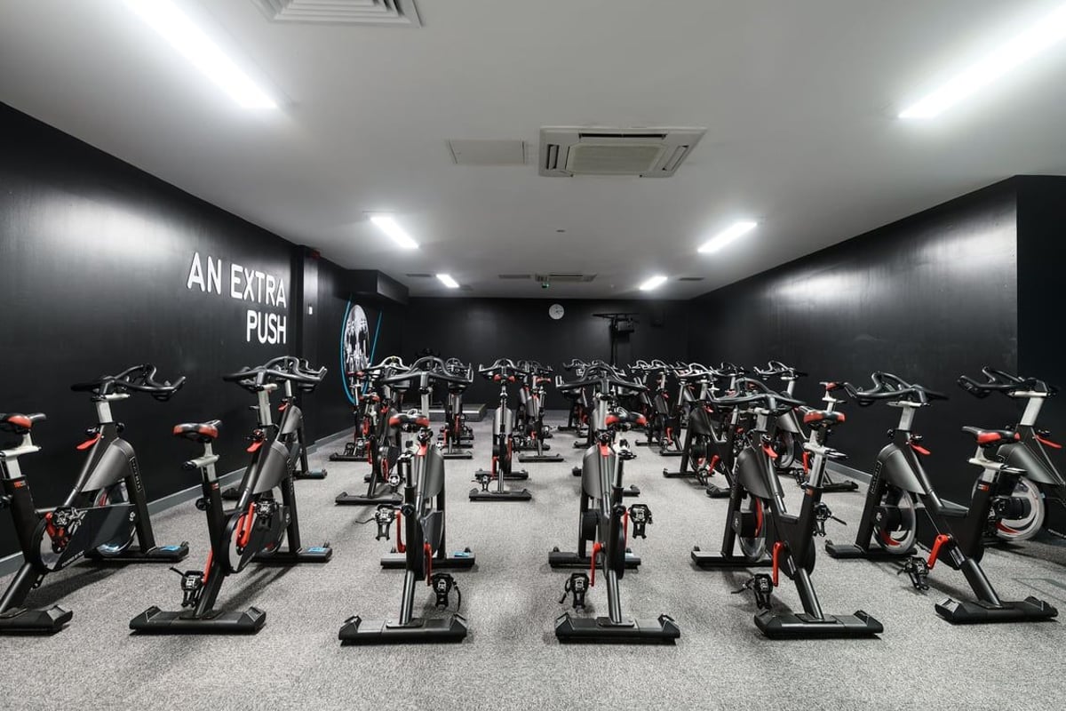 PureGym Newcastle Longbenton: Rows of stationary exercise bikes in a dimly lit studio with black walls and neon signage.