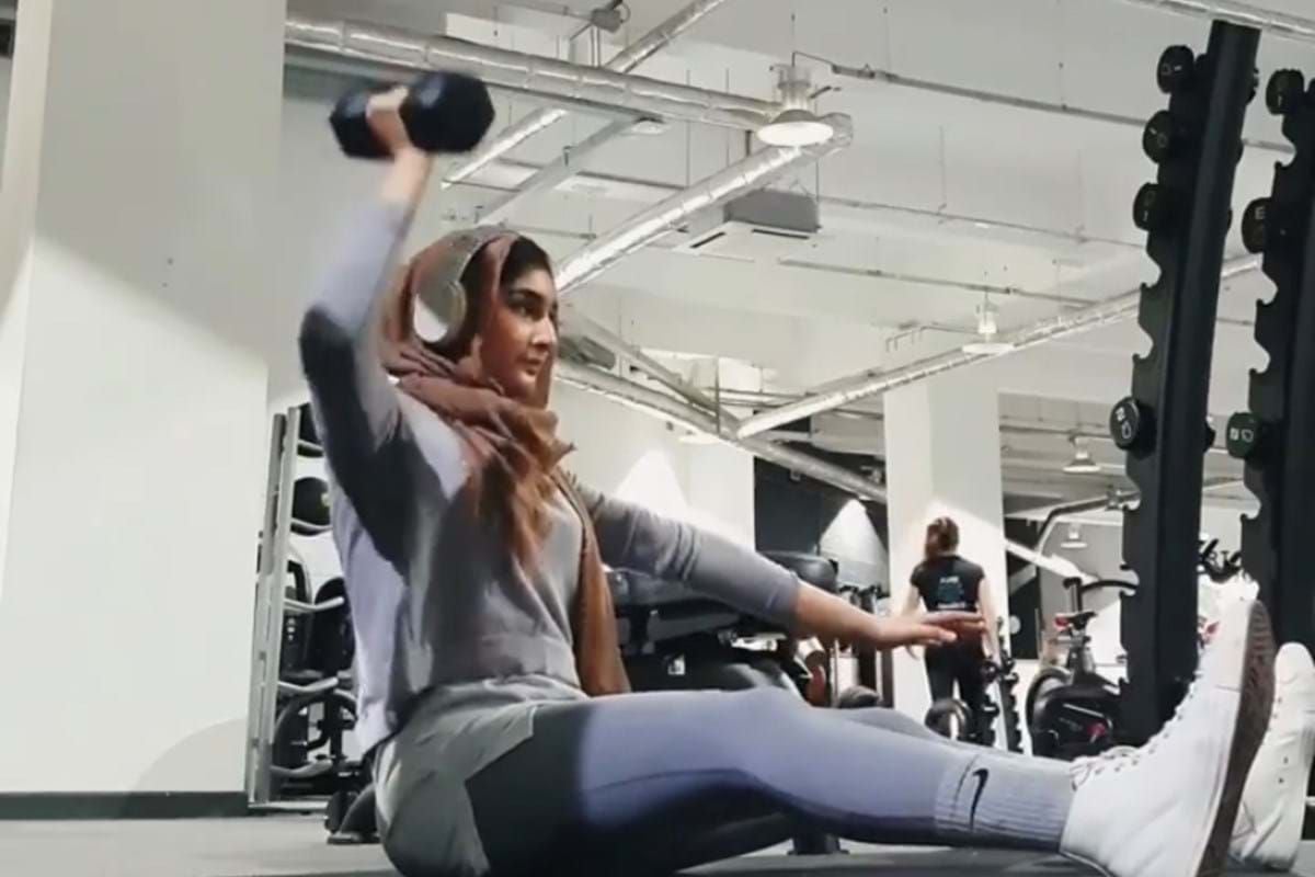 Woman in hijab lifting dumbbell while seated on gym floor with legs extended