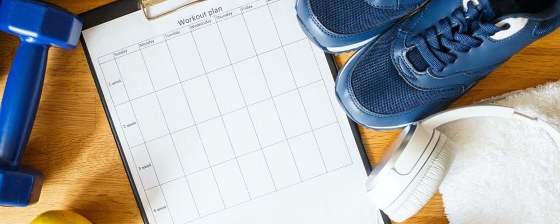 Workout plan sheet with blue trainers, dumbbell, apple, headphones, and towel on wood