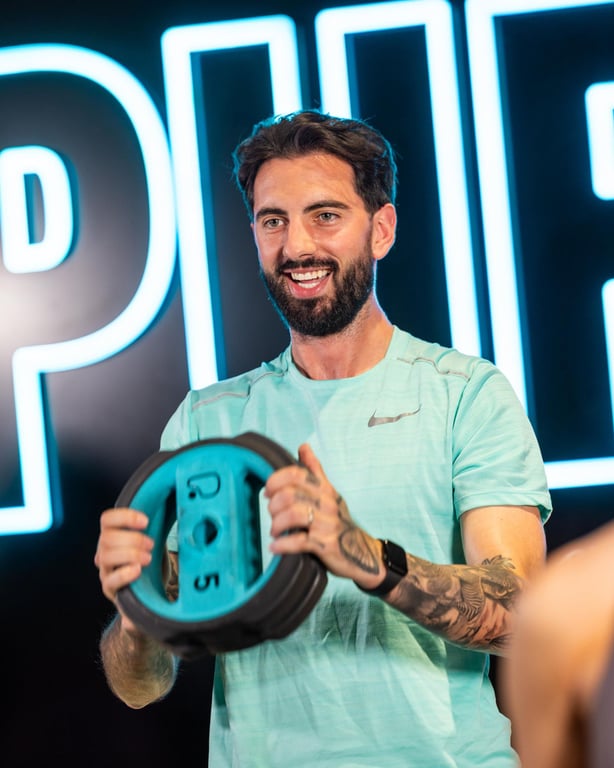 Lächelnder Mann mit Tattoos im türkisfarbenen Shirt hält eine türkisfarbene Hantelscheibe vor dem PureGym-Neonschild