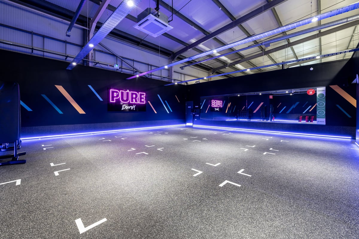 PureGym Swindon Stratton: Indoor pure cycling studio with neon lighting, rubberized flooring, and marked cycle positions.