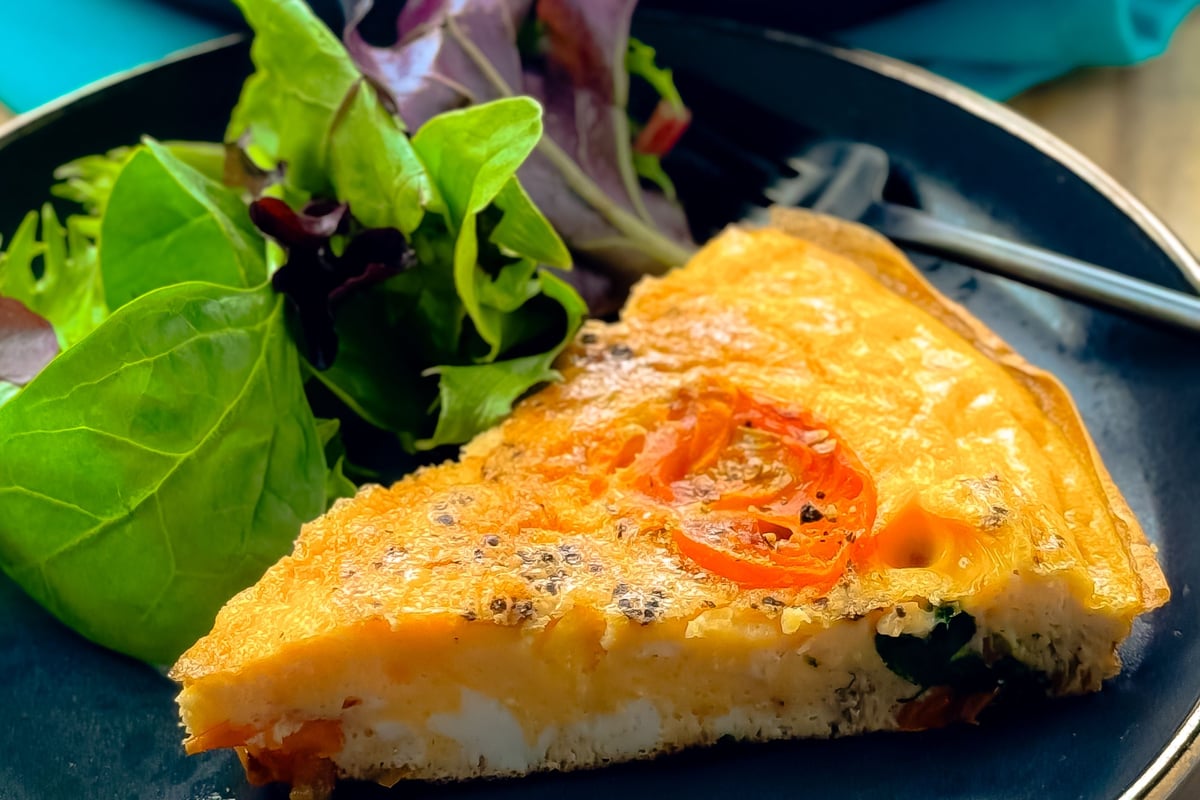 Slice of vegetable frittata with salad greens on a dark plate with a fork