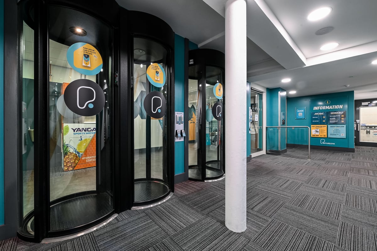 PureGym London Piccadilly: Revolving entrance with promotional posters, digital screens, and teal-coloured decor.