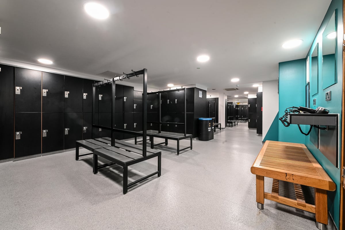 PureGym London Piccadilly: Rows of lockers, benches, and a woodgrain counter in a locker room setting.