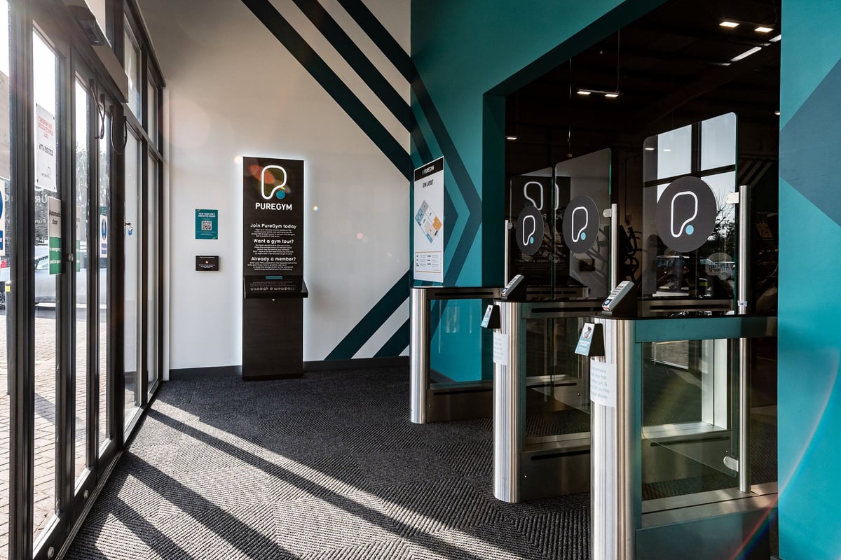 PureGym Newbury: Gym entrance with revolving security gates, digital kiosks, and striped design elements.