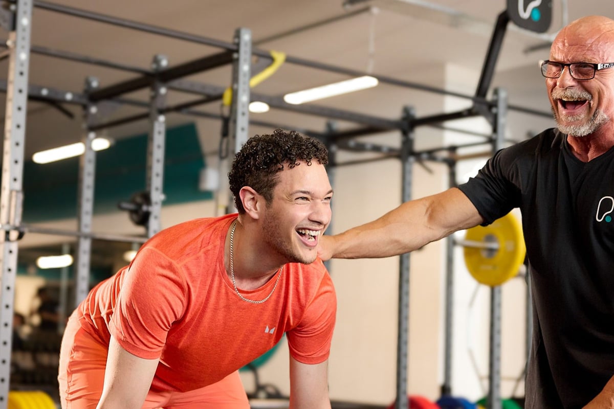 Young man in orange shirt laughing with older man in black shirt at gym