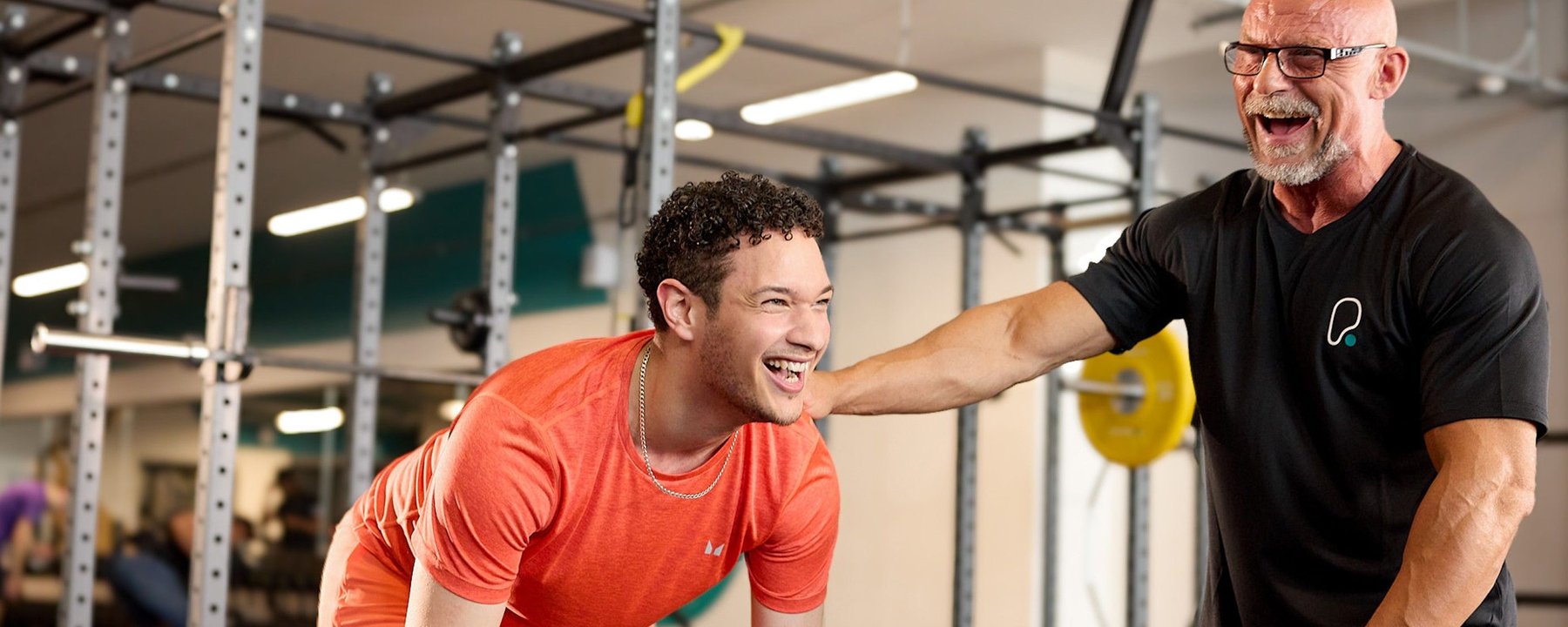 Young man in orange shirt laughing with older man in black shirt at gym