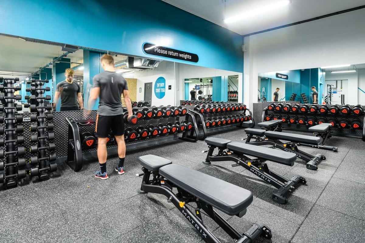 PureGym East Grinstead: Rows of dumbbells, benches, and rubberized flooring in a blue-tinted gym with two people visible.