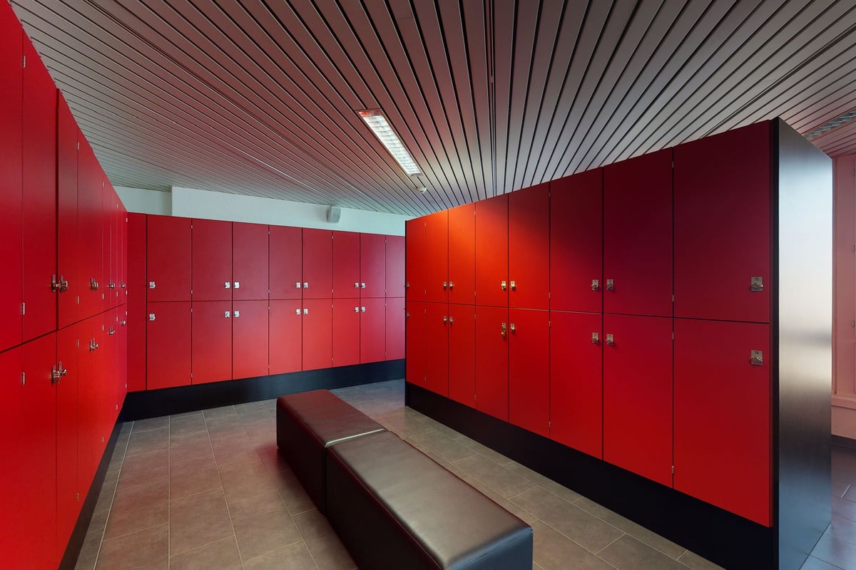 PureGym Kriens: Red locker room with benches in the centre, featuring rows of lockers along the walls and slatted ceiling.