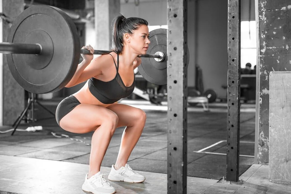 Frau führt Kniebeugen mit Langhantel im Fitnessstudio durch, Gewichte und Metallgestelle s