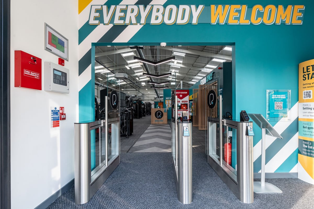 PureGym Keighley: Corridor with turnstiles, signs indicating welcome, and flags.