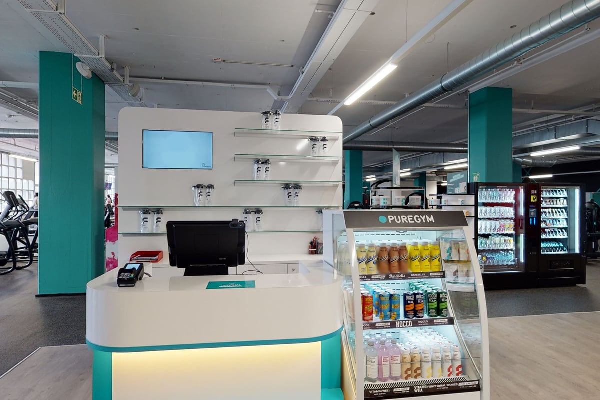PureGym Bern Bümpliz: Hall d'entrée avec distributeurs automatiques, comptoir de service et présentoirs électroniques présentant les produits.