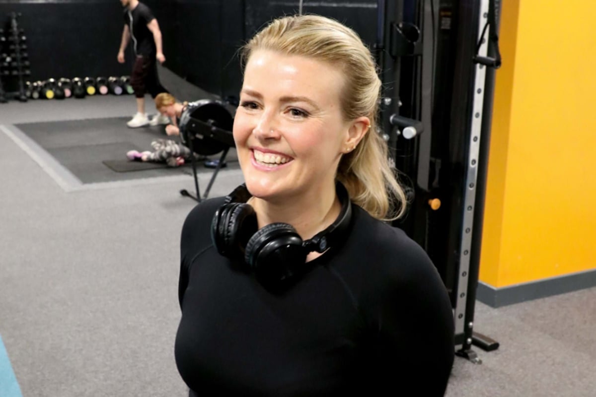 Smiling woman with headphones around her neck in a gym with people exercising behind her