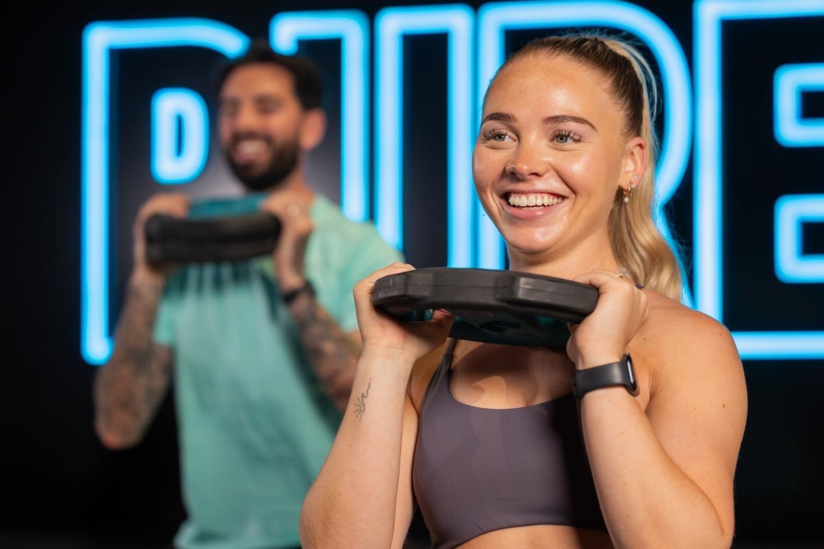 Woman in gym holding weight plate at shoulder height with man exercising behind her