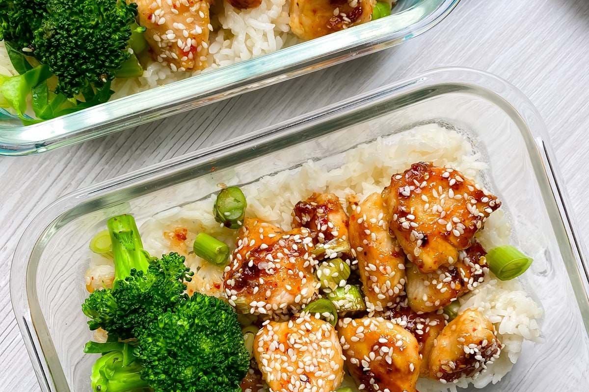 Two glass containers with rice, glazed chicken pieces, broccoli, and sesame seeds on a tab